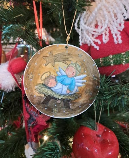 Adorno redondo colgado en árbol navideño con ángel junto al Niño Jesús bajo estrella fugaz