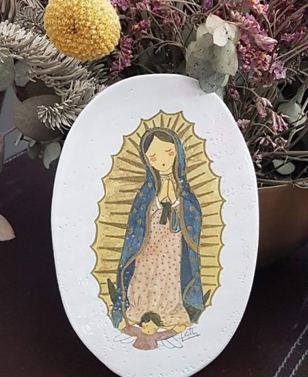 Medalla de cerámica con imagen de la Virgen de Guadalupe, colocada sobre mesa con flores secas